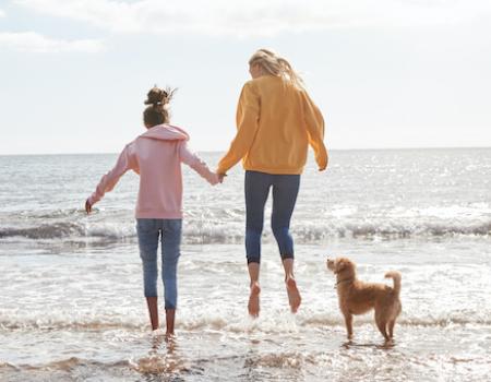 mom and daughter on beach with dog jumping in the water with jeans and sweaters on