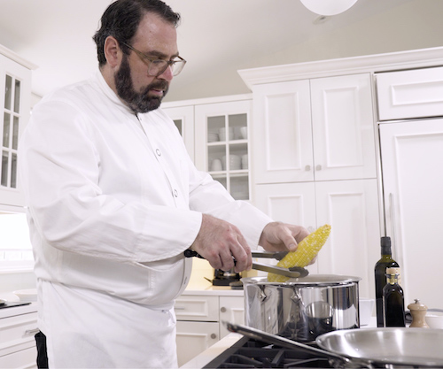 chef in white coat cooking corn on stove in all white kitchen