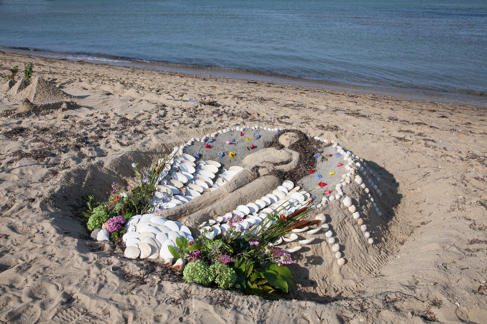 sandcastle art with woman carved out, shells, and other materials circling the image